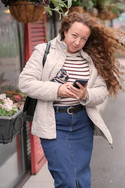 Kıvırcık saçlı bir kadın tuhaf bir sokakta yürüyor, çiçeklerin yanındaki telefonunu kontrol ediyor..