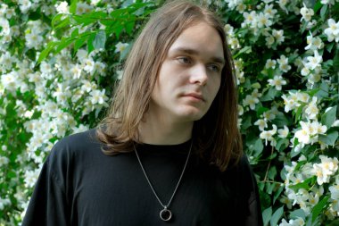 Uzun saçlı genç bir adam, titreyen beyaz çiçeklerin arasında sessizce duruyor, sakince yayılıyor..