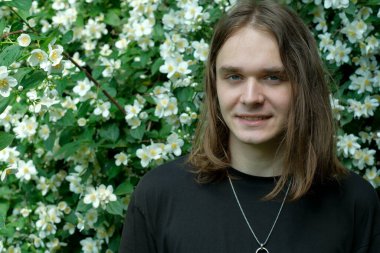 Uzun kahverengi saçlı genç bir adam çiçeklerle dolu bir bahçede gülümsüyor..