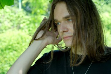 Dalgın, uzun saçlı genç bir adam canlı bir yeşilliğin ortasında duruyor, düşüncelere dalmış..