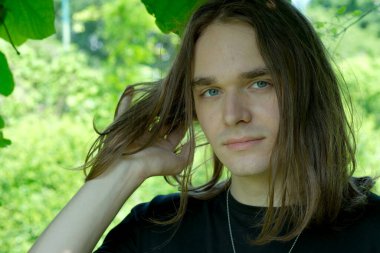 Genç bir adam yemyeşil bir ortamda duruyor, sakin bir neşeyle saçlarına dokunuyor..