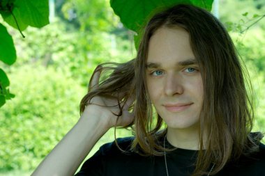 Dalgalı saçlı genç bir adam zarif yeşil bir ortamda poz verir..