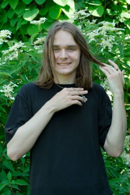 Neşeli genç bir adam canlı bir bahçede uzun saçlarına hafifçe dokunur..