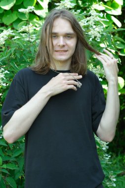 Genç bir adam yemyeşil bir yerde duruyor, nazikçe saçlarına dokunuyor ve gülümsüyor..