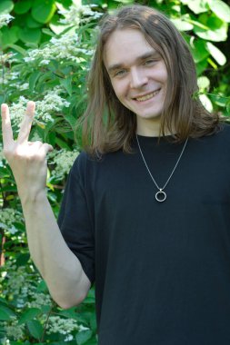 Neşeli bir birey canlı bir yeşilliğin ortasında duruyor, mutluluk içinde bir barış işareti gösteriyor..