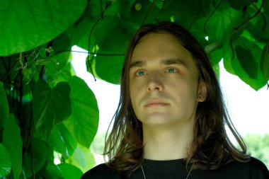 Uzun saçlı genç bir adam canlı yeşil yaprakların arasında düşünceli bir şekilde duruyor..