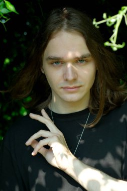 Uzun saçlı genç bir adam yemyeşil ve gölgelerin arasında duruyor..