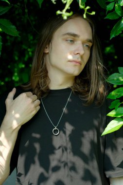 Genç bir adam sessizce duruyor, parlak yeşil yaprakların arasında dalgın dalgın..