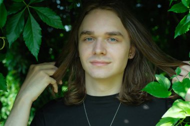 Genç bir adam uzun saçlarını canlı yeşil bir ortamda nazikçe tutarken gülümsüyor..