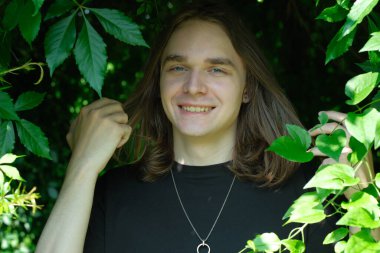 Neşeli genç bir adam canlı yeşil yaprakların arasında saçlarına neşeyle dokunuyor..