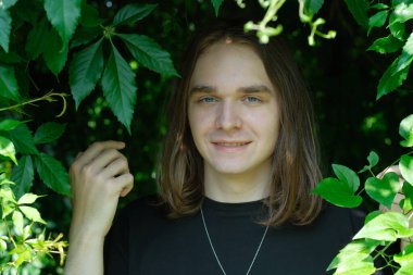 Genç bir adam canlı yeşil yapraklarla çevrili duruyor, doğada parlak bir şekilde gülümsüyor..