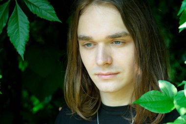 Uzun saçlı genç bir adam canlı yapraklarla çevriliyken düşünceli bir şekilde bakar..