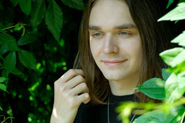 Uzun saçlı genç bir adam çevrende canlı yeşil yapraklar varken hafifçe gülümsüyor..