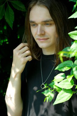 Genç bir adam yeşil yaprakların arasında duruyor, güneş ışığı yüzünü aydınlatıyor..