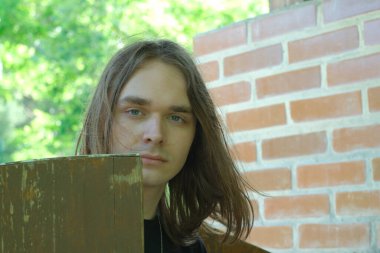 Dalgalı saçlı genç bir adam düşünceli bir şekilde kırsal bir ahşap çitin yanında duruyor..