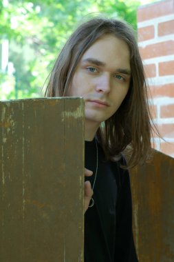 Çarpıcı gözleri ve uzun saçları olan genç bir adam tahtanın içinden düşünceli görünüyor..