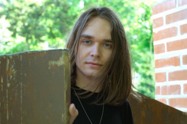 Genç bir adam gün ışığında gülümserken ahşap bir yapıya yaslanır..