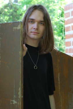 Uzun saçlı genç bir adam yıpranmış ahşap bir çitin yanında duruyor, huzur saçıyor..