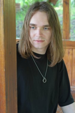 Uzun saçlı genç bir adam sessizce bir kulübede duruyor, yansımasında kaybolmuş..