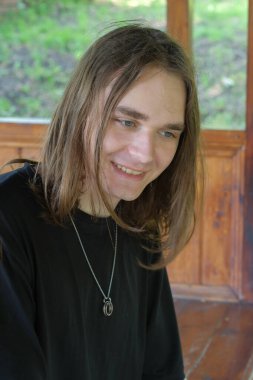 Uzun saçlı neşeli bir genç adam dışarıda eğleniyor, sıcak bir şekilde gülümsüyor..