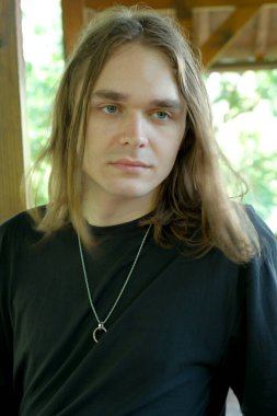 Uzun saçlı genç bir adam güneşli bir alanda dikiliyor, düşünceli bir şekilde ileriye bakıyor..