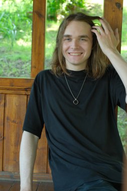 Uzun saçlı genç bir adam, doğayla çevrili ahşap bir çardakta rahatlarken gülümsüyor..