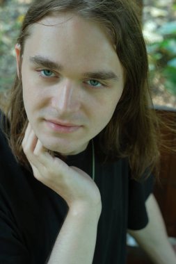 Dalgalı saçları ve çarpıcı mavi gözleri olan genç bir adam sakin bir açık hava ortamında dinleniyor..