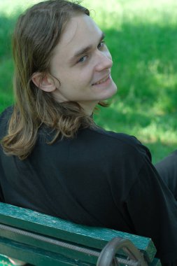 Uzun saçlı genç adam yeşil ağaçların altındaki bankta rahatlar, sıcak bir şekilde gülümser..