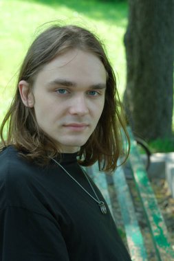 Uzun saçlı genç bir adam parktaki bankta oturur, güneşli bir günde.