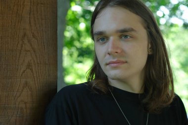 Genç bir adam düşünceli bir şekilde yeşilliklerle çevrili ahşap bir sütunun yanında duruyor..