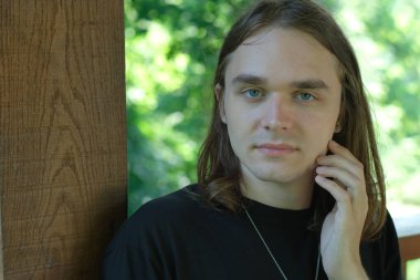 Genç bir birey ahşap bir yapının yanında duruyor, yeşillikler arasında kaybolmuş..