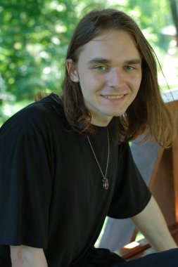 Uzun saçlı genç bir adam yeşilliğin ortasında dinlenirken sıcacık gülümsüyor..