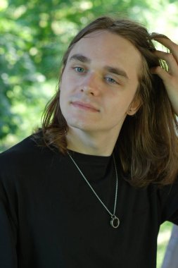 Uzun saçlı genç bir adam yemyeşil bir ortamda duraklıyor, düşüncelere dalmış..