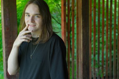 Düşünceli genç bir adam, yemyeşil yemyeşil ağaçlarla çevrili ahşap bir yapının altında duruyor..