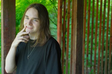 Genç bir adam düşünceli bir şekilde yeşilliklerle çevrili ahşap bir yapının yanında duruyor..