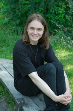 Genç bir adam, canlı yeşillikler ve çiçeklerin ortasında, ahşap bir bankta dinleniyor..