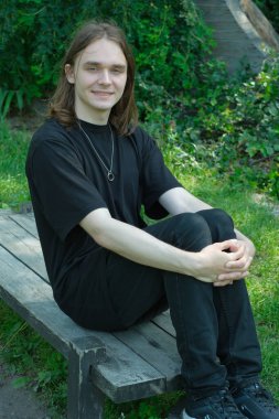 Genç bir adam canlı yeşilliklerle çevrili bir bankta rahatça oturuyor..