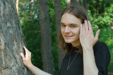 Genç bir adam bir ağacın yanında duruyor, sakince gülümsüyor ormanın arka planında..