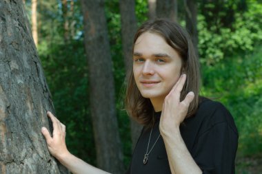 Genç bir birey yumuşak bir şekilde uzun bir ağacın yanında duruyor, sakin orman çevresinin tadını çıkarıyor..