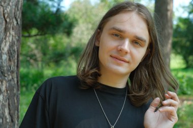 Genç bir insan bir ağacın yanında duruyor, dışarıda dinlenirken nazikçe gülümsüyor..