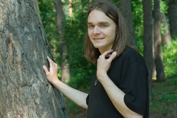 Bir insan, yemyeşil yemyeşil bir ağaçla çevrili, uzun bir ağacın yanında duruyor, yavaşça gülümsüyor..