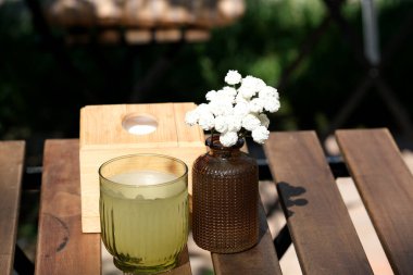 Bir bardak limon içeceği, parlak güneş ışığı altında küçük beyaz çiçeklerden oluşan bir vazonun yanında duruyor..