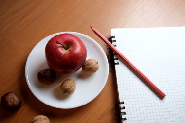 A bright red apple with walnuts sits on a plate near a notepad and pencil.