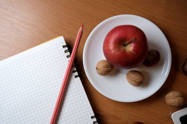 A red apple and walnuts sit on a white plate next to a notebook and pencil.