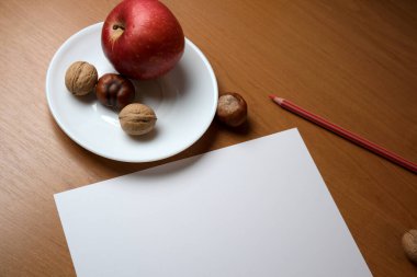 Fresh apple and assorted nuts sit on a plate beside blank paper and a pencil.