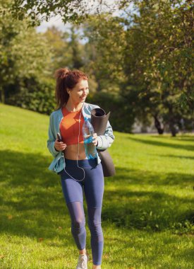  Renkli fitness kıyafetli beyaz bir kadın yoga minderi şişesi ve akıllı telefonuyla parkta yürüyor.