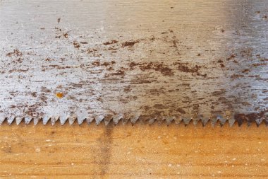 Sıcak ahşap bir tahtanın üzerinde rustik bir el testeresi bıçağının yoğun yakın çekimi. Çizik metal, pas ve ahşap tahıl dokuları marangozluk, DIY projeleri ve işçilik kavramlarını çağrıştırıyor. El emeği ve yaratılış temaları için ideal arkaplan.