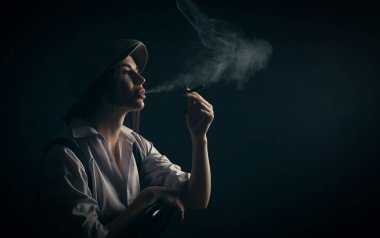 Genç bir kadın karanlık bir arka planda oturup sigara içiyor.