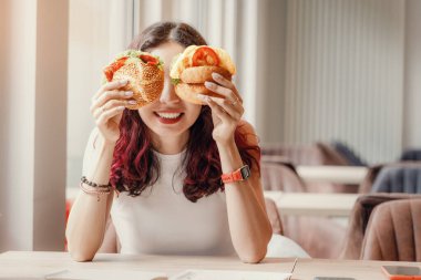 Bir kafede oynaşan ve gözlerini iki çizburgerle kapatan bir kız. Fast food ve sağlıklı diyet konsepti