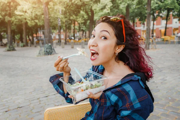 A happy girl is snacking on a ready-to-eat salad in a city park. Diet and cheap takeaway fast food on the street